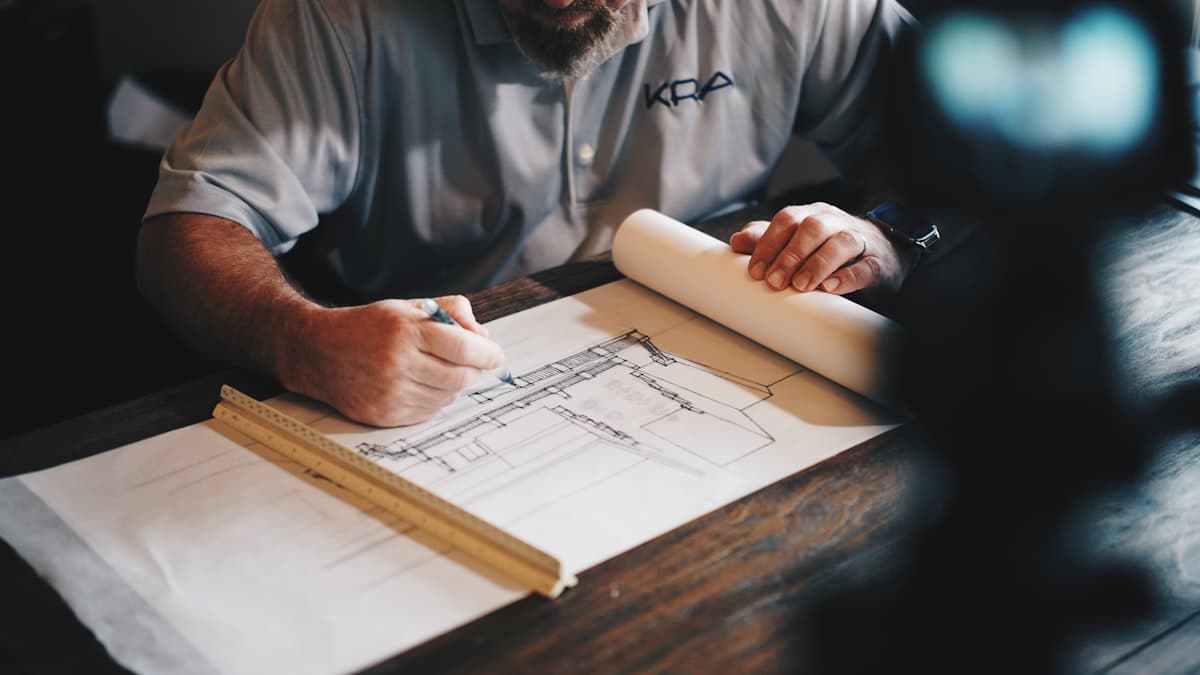 Architect drafting a detailed construction drawing with a scale ruler and rolled plans on a wood desk